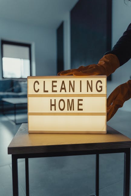 A person wearing orange gloves is placing a illuminated sign on a wooden table that reads 'CLEANING HOME', indicating home cleaning services. The background features a modern interior with blue walls,