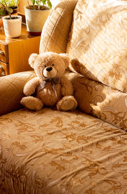 A person using a handheld vacuum cleaner to deep clean a decorative yellow and white patterned cushion on a dark fabric sofa in a living room, with natural lighting highlighting the surface's cleanlin