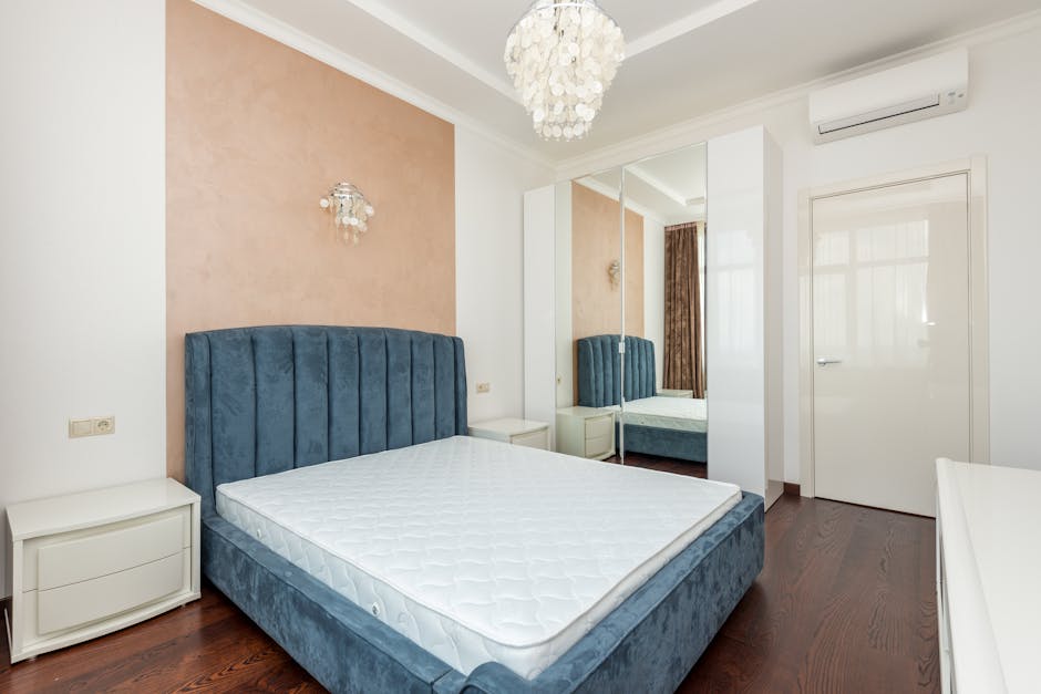 A modern bedroom featuring a large, light-colored upholstered bed with a thick mattress, flanked by matching dark wooden bedside tables equipped with white ceramic table lamps. The room has beige wall